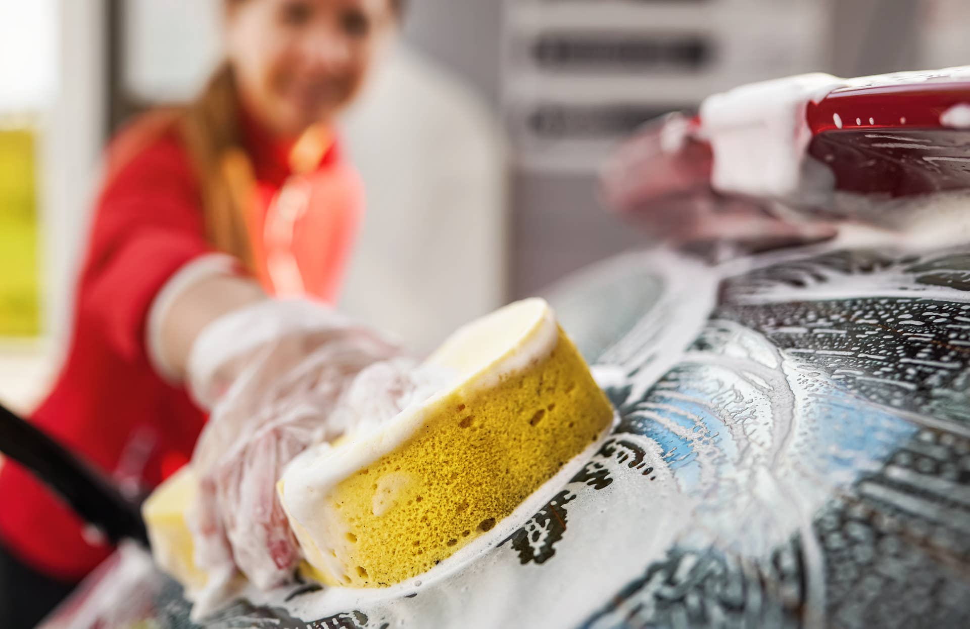 Young woman cleaning rear window of her car, closeup detail on hand in glove holding yellow sponge
