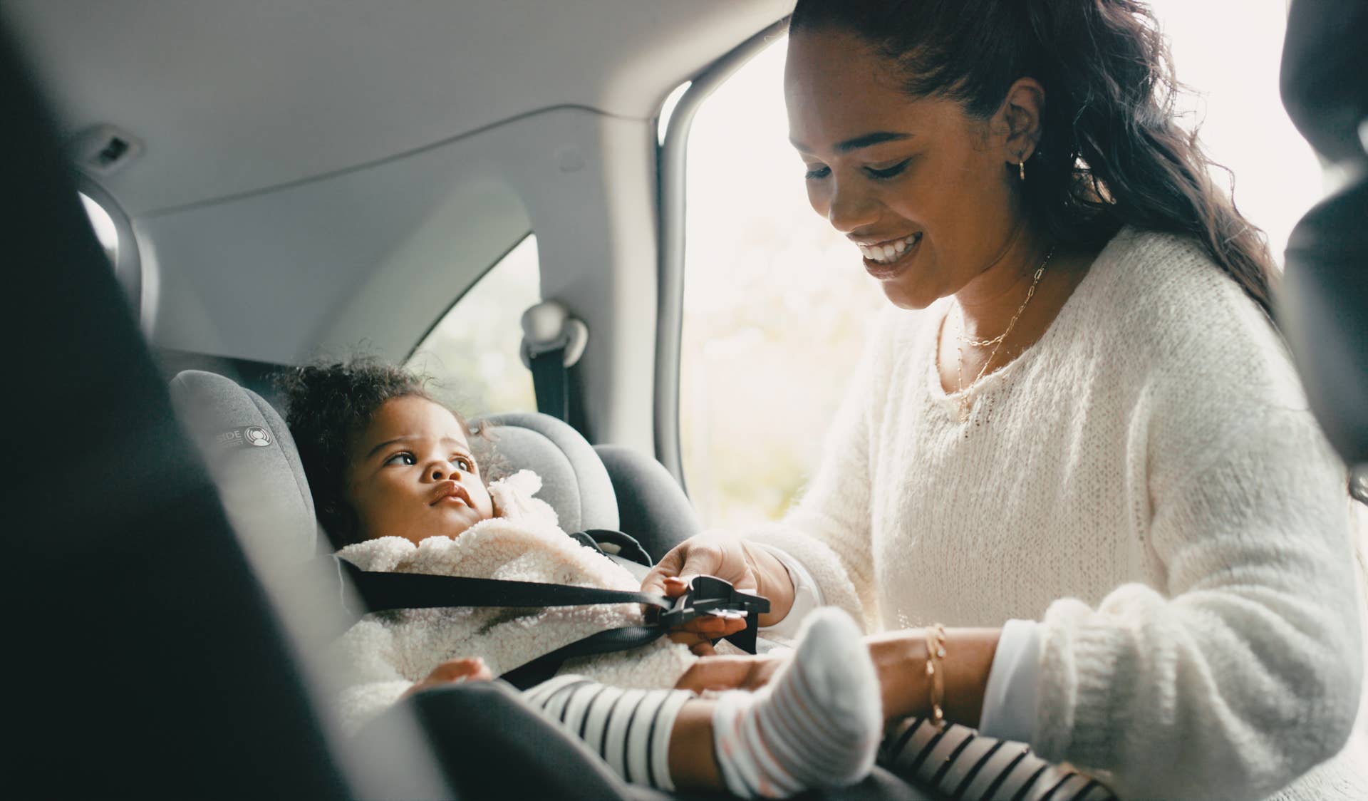 Happy, mother and baby in a car seat for safety, security and support during a drive in car. Happiness, smile and young woman secure her girl child into infant seat with care to travel with vehicle.