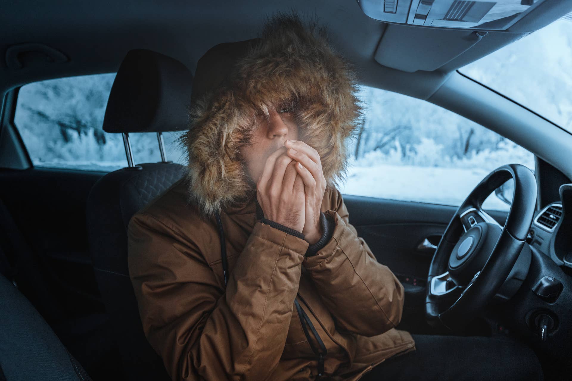 Man in down jacket tries to stay warm in stalled car during winter.