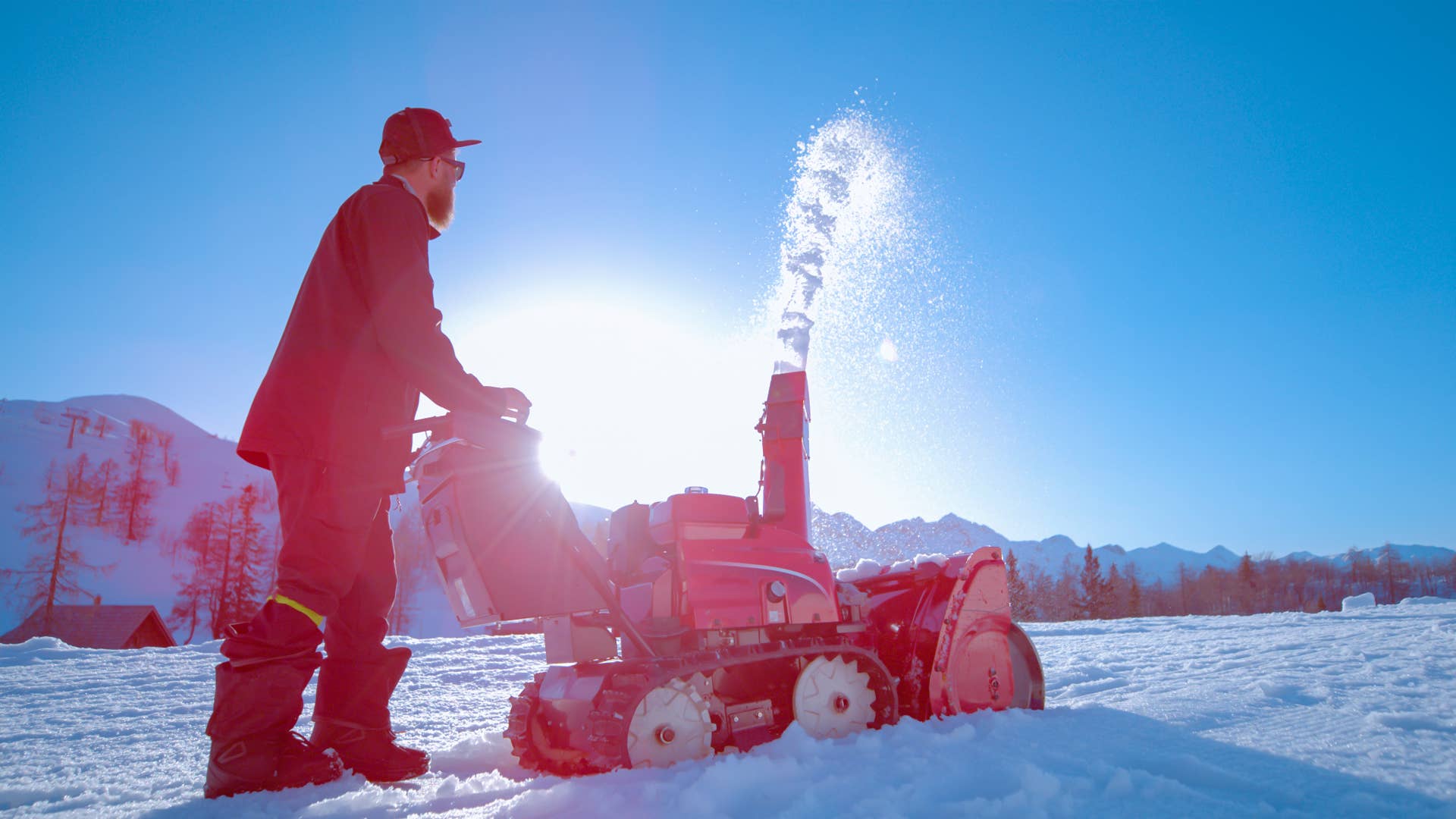 LOW ANGLE, LENS FLARE: Bright sunbeams shine on a bearded male shaper using a snow blower to create a kicker out of fresh snow. Industrial machinery operator blows snow into air on sunny winter day.