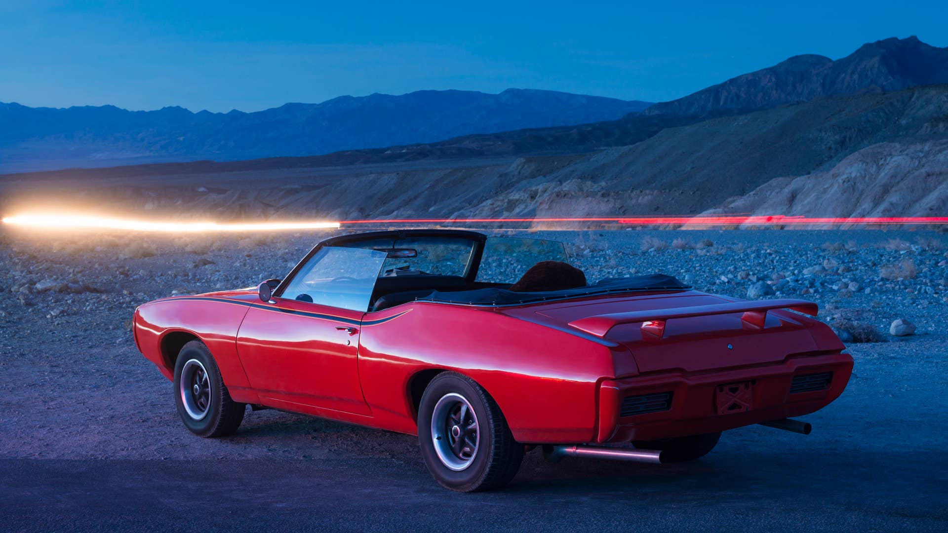 A red 1969 vintage Pontiac GTO Judge convertible open top car parked in Death Valley near Zabriske Point before distant mountains and light trails from other cars speeding past