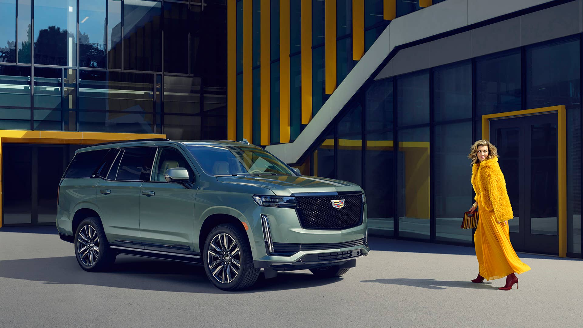 Front 3/4 view of a 2024 Cadillac Escalade Sport in Dark Emerald in front of a building with a woman standing next to it.