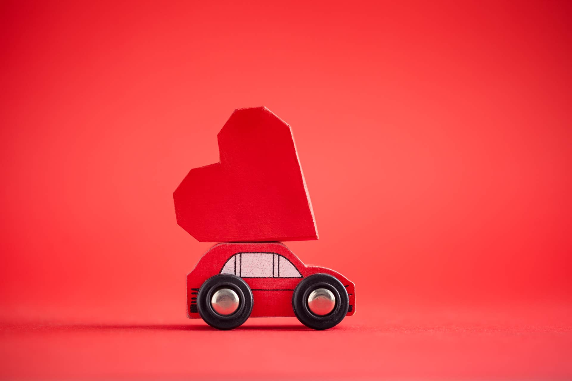Red model of car delivering red paper heart on red background. February 14 card for holiday concept. Creative greeting card for Saint Valentine day.