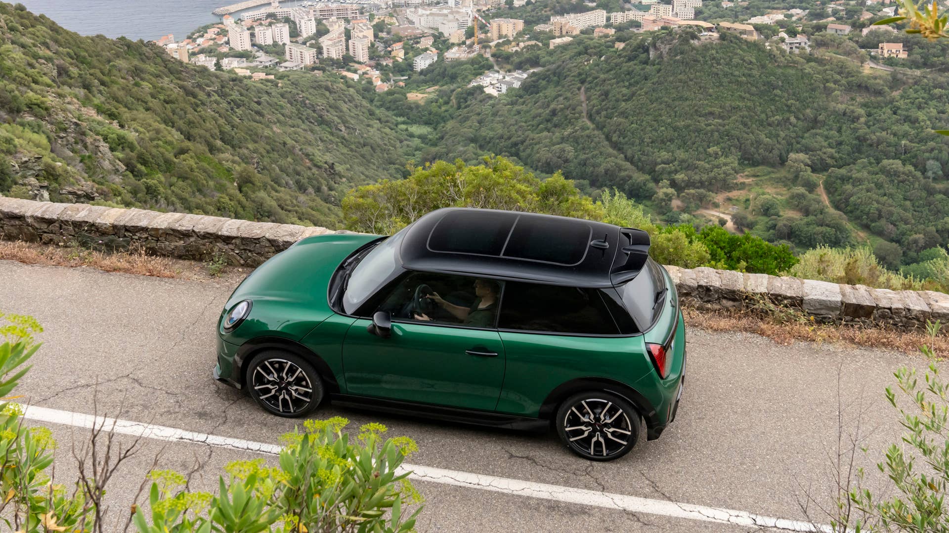 Green 2025 Mini Cooper S JCW driving on a mountain road, top side view.