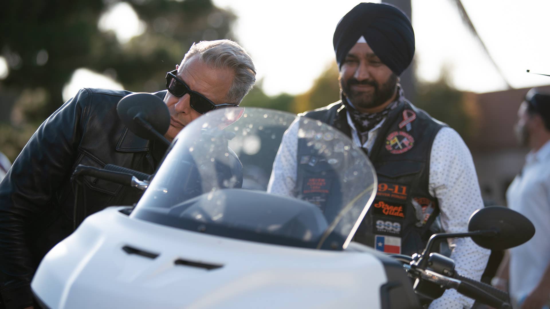 Huntington Beach, CA - Jeff Goldblum (L) looking in the wing mirror of a motorcycle as a motorcycle enthusiast stands by. (Credit: National Geographic)