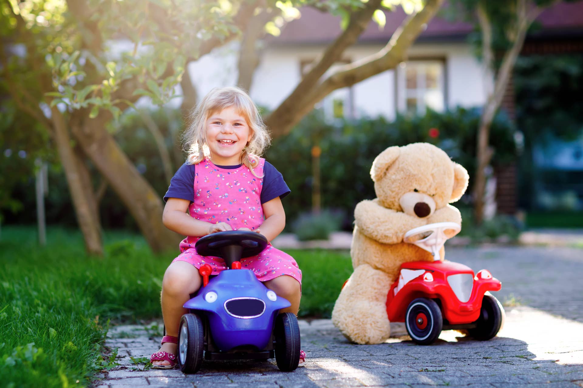 Little adorable toddler girl driving toy car and having fun with playing with plush toy bear, outdoors. Gorgeous happy healthy child enjoying warm summer day. Smiling stunning kid in gaden.