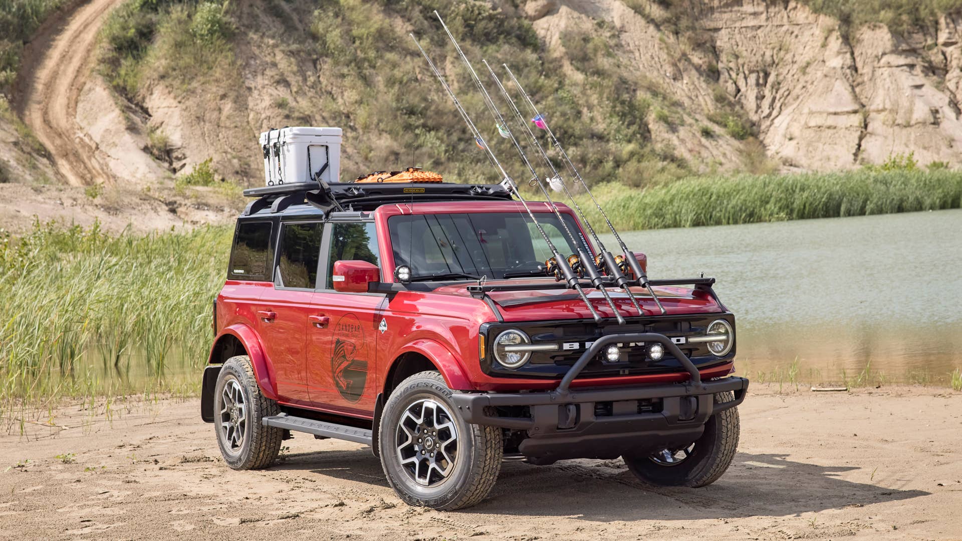 Bronco Four-Door Outer Banks Fishing Guide. Purpose-built to support a professional fishing guide along Cape Hatteras National Seashore, this lifestyle concept is based off the regionally inspired four-door Bronco Outer Banks™ series. On top is a Bestop® Sunrider® first-row soft top, factory-style concept roof rails and crossbars, and a Yakima® LockNLoad™ Platform roof rack. The SUV makes use of fender-mounted trail sights to fit a custom-made fishing pole and seat perch to wait for the perfect catch. A Ford Performance modular front bumper and safari bar help push through rugged terrain, while a slide-out tailgate* provides a great work surface when repairing fishing rods or stringing up new lures. All-weather floor mats and splash guards round out the package. (Preproduction image with optional features shown. Available beginning Spring 2021.) (*Available late 2021)