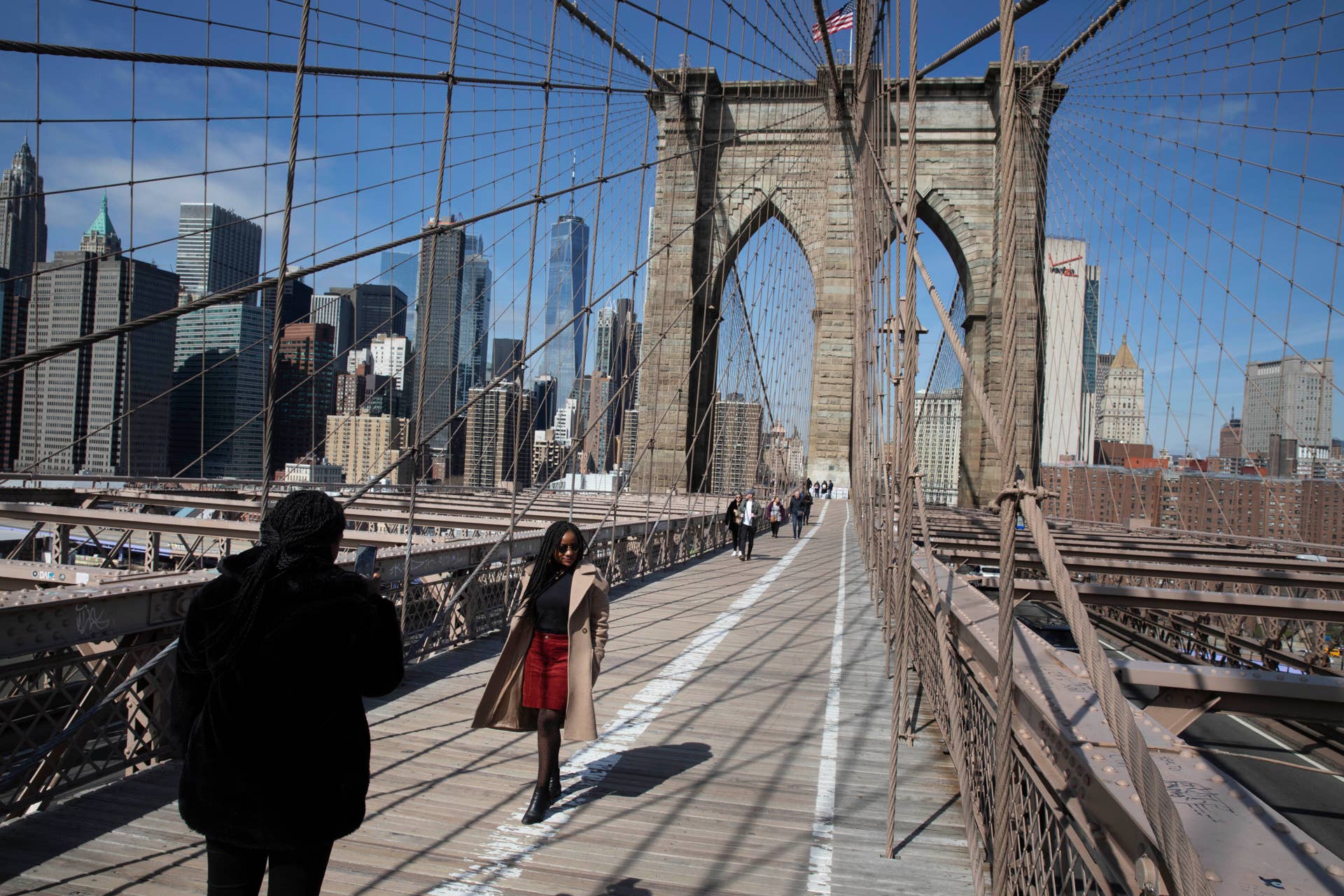 Ines Tshiyomba, center, poses as her friend Garethe Mawonso takes her photo on the Brooklyn Bridge, Monday, March 16, 2020 in New York. The Parisians are stuck in New York for another day as their return flight was cancelled. The bridge's pedestrian path is normally crowded on a sunny day. (AP Photo/Mark Lennihan)