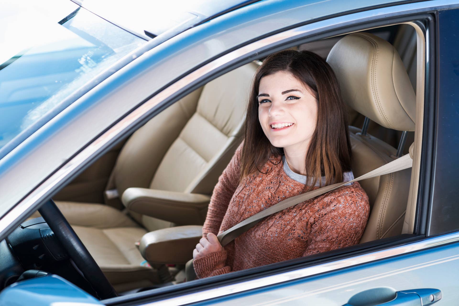 A teenage girl driving a car. She is sitting in the driver's seat, buckling her seat belt, smiling confidently at the camera through the open window. She is learning to drive, or maybe just got her new license.