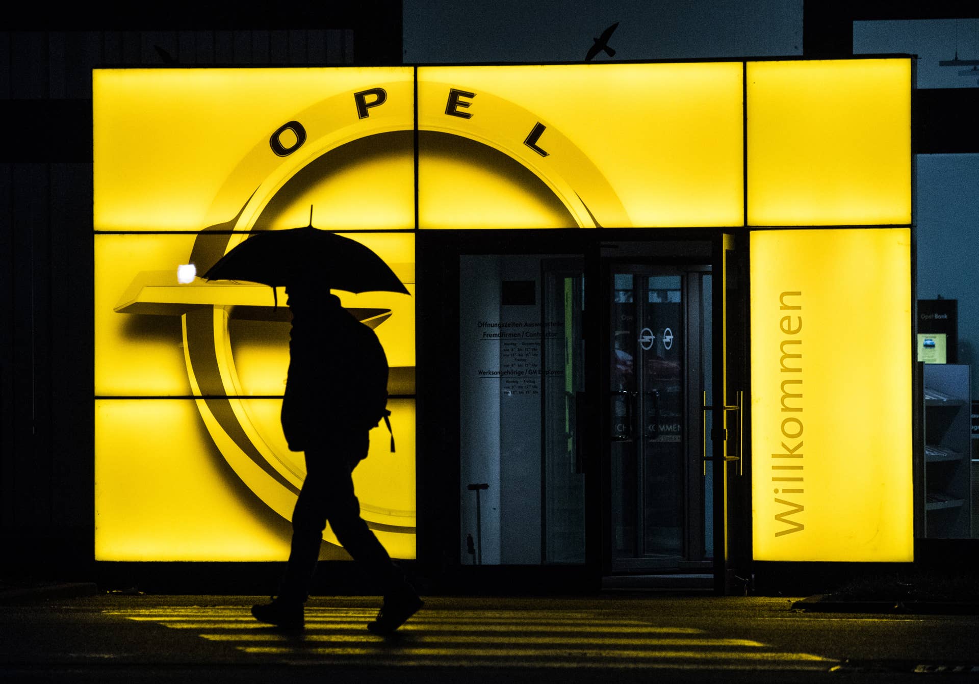 A pedestrian shelters under an umbrella while passing an illuminated Adam Opel AG logo outside the Opel automobile factory in Ruesselsheim, Germany, on Thursday, Nov. 9, 2017. PSA Group�will offer electric and hybrid versions across the entire Opel lineup by 2024, tap overseas markets and cut vehicle costs at its new unit after acquiring the German carmaker from�General Motors Co.�earlier this year. Photographer: Andreas Arnold/Bloomberg via Getty Images
