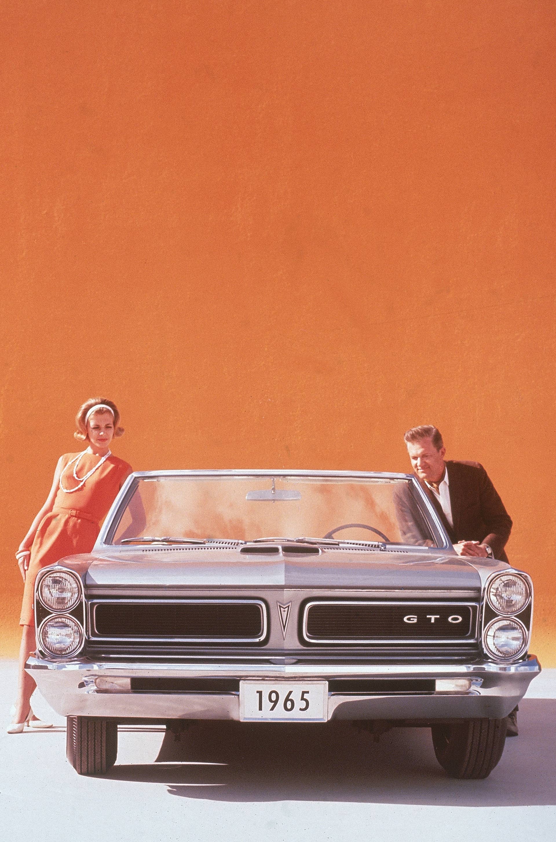 1965: A couple posing on either side of a silver 1965 Pontiac GTO convertible sports car in front of an orange backdrop. (Photo by Hulton Archive/Getty Images)