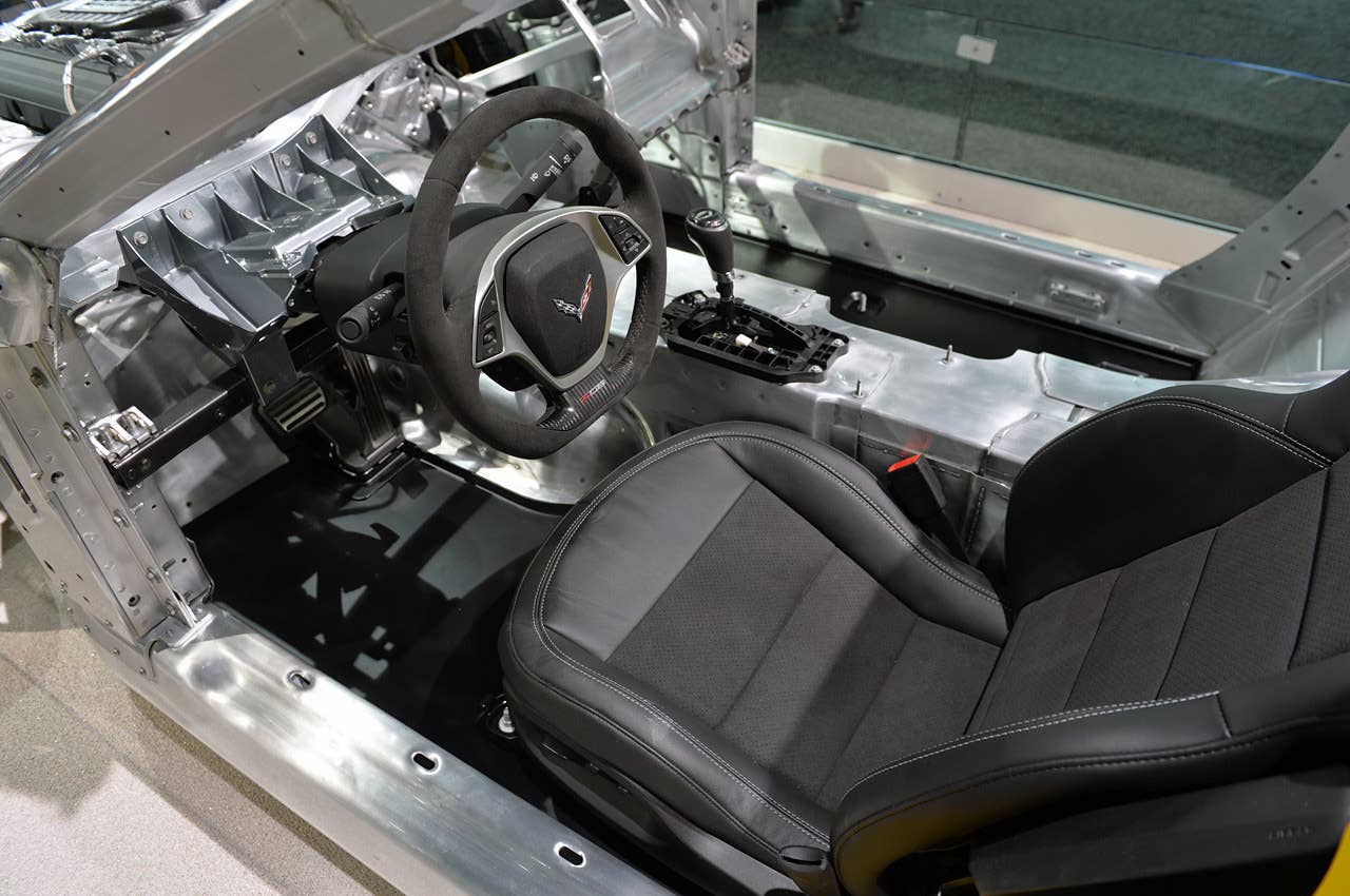 This is a clean shot of the driver's compartment, with the finished seat, steering wheel and shifter in place. A boxed steel tube runs beneath the steering column, bolted to the vertical A-pillar on the left of the image. Note the substantial bracket holding the microsuede wheel in place.