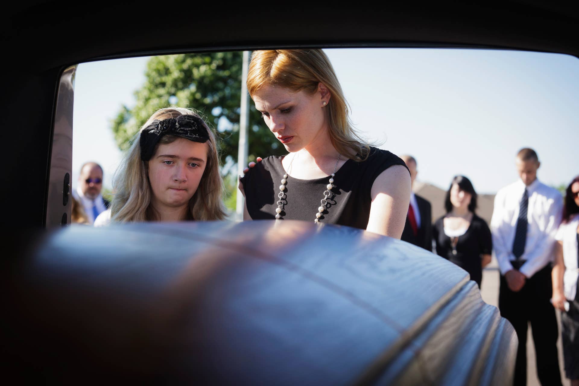 A grieving woman standing at the rear of a hearse looking down at the casket. Taken from within the hearse.