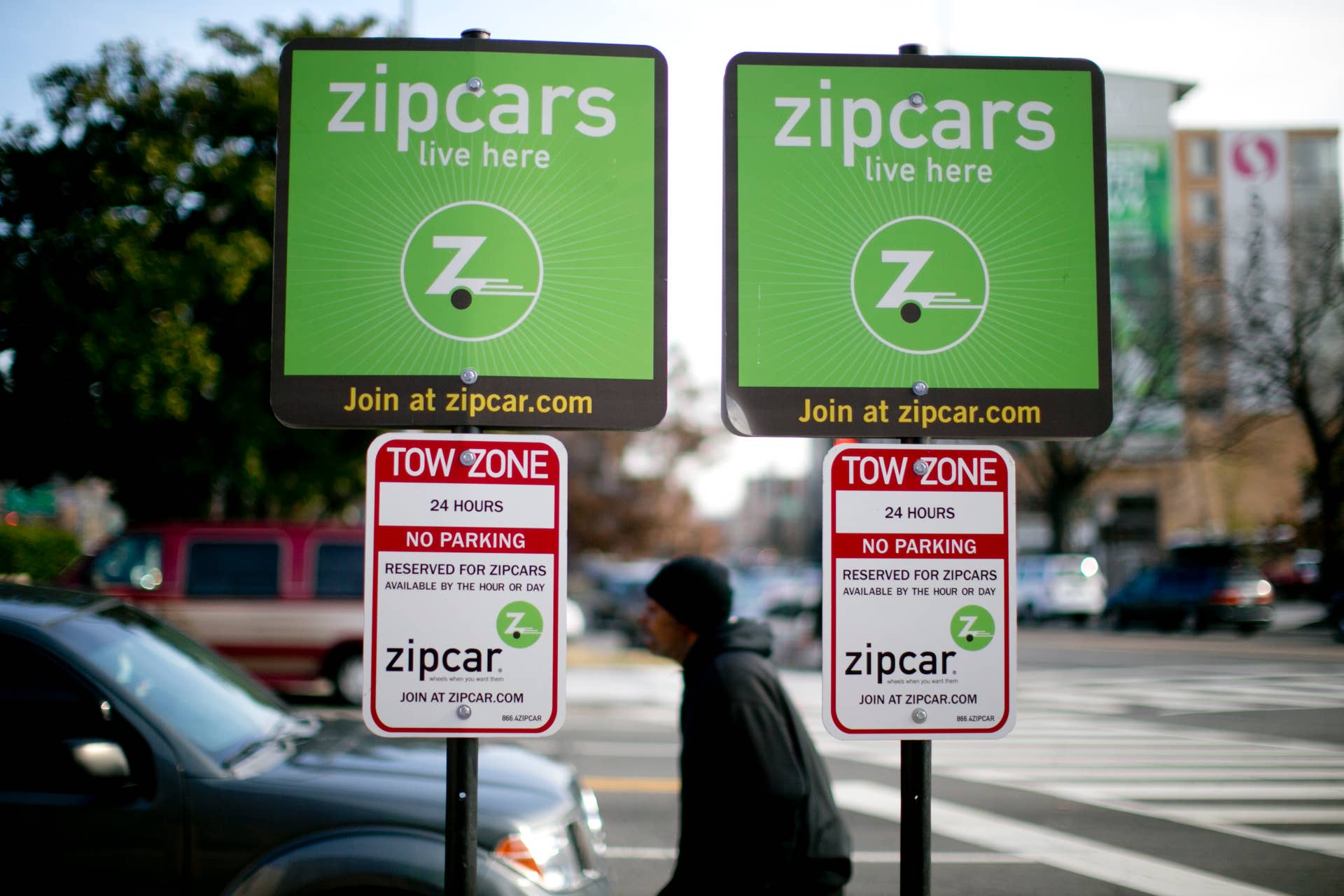 A pedestrian walks past Zipcar Inc. signs standing in the company's parking spaces in Washington, D.C., U.S., on Wednesday, Jan. 2, 2013. Avis Budget Group Inc., once a skeptic about car sharing services, agreed to buy short-term rental pioneer Zipcar for $491 million, signaling a shift in the industry to embracing drivers who donât want to own cars. Photographer: Andrew Harrer/Bloomberg via Getty Images