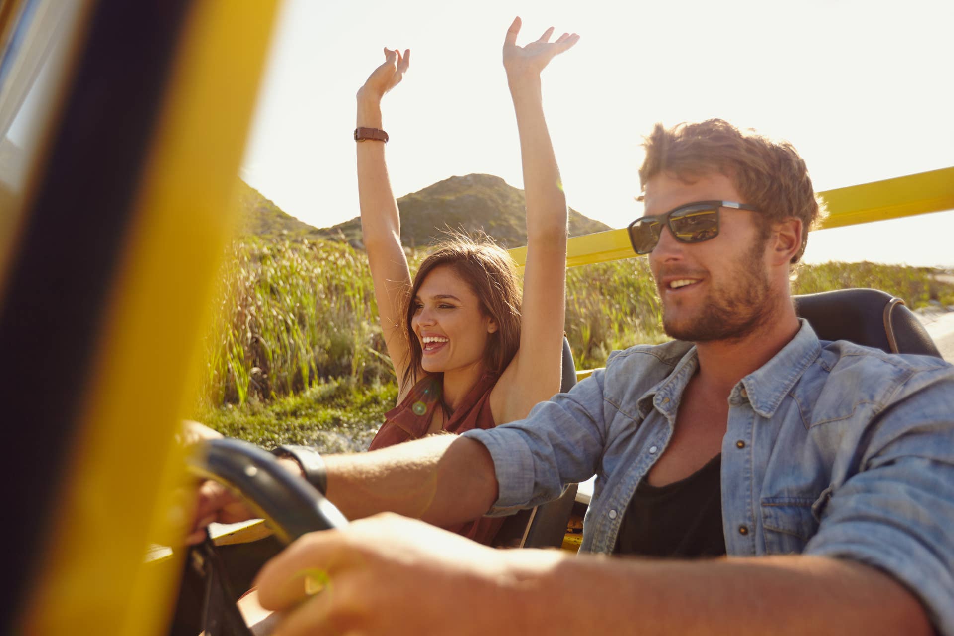 Happy couple enjoying on a long drive in a car. Friends going on road trip on summer day. Caucasian young man driving a car and joyful woman with her arms raised.