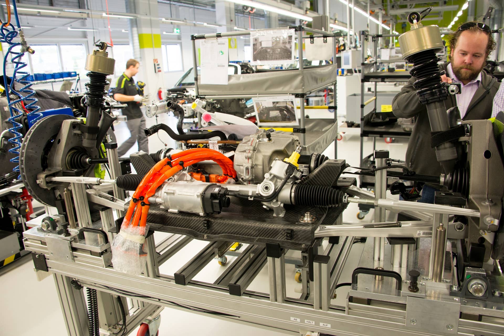 Loaded on a wheeled dolly, the front clip, complete with electric drive motor, steering rack, suspension, hubs and brakes, waits to be assembled into the nose of a 918. After inspection, it will be rolled over the tub and lifted into place. The heavy orange cables will be connected to the high-voltage electric system, while the steering column will bolt onto the yellow fitting (seen near the middle of the image).