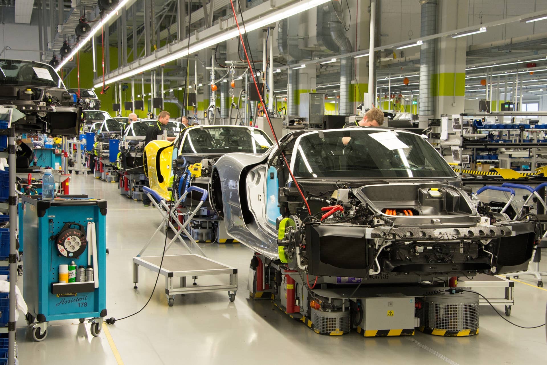 Porsche assembles all 918 Spyder models in Zuffenhausen, Germany, in a building adjoined to the 911 production line. The large well-lit room used to be a dedicated 911 paint shop, but it was re-purposed and completely remodeled with white walls and floors, accented with Porsche's signature "hybrid" Acid Green paint.  The room is remarkably quiet, lacking the clatter and crashing of pneumatic power tools and stamping equipment. Instead, there is only the whir of Bluetooth-enabled battery operated electric hand tools that keep a digital record of all of their work. That silence carries through the production— the 918 models, when completed, drive out of the assembly hall under silent electric power.   There are 110 assembly workers in the room, both men and women, who are assigned to one of 15 different assembly stations. The line remains absolutely motionless for 111 minutes before moving forward one station— usually just a few feet away. The work is slow and meticulous — on a typical day, just four new 918 Spyders will leave the factory.