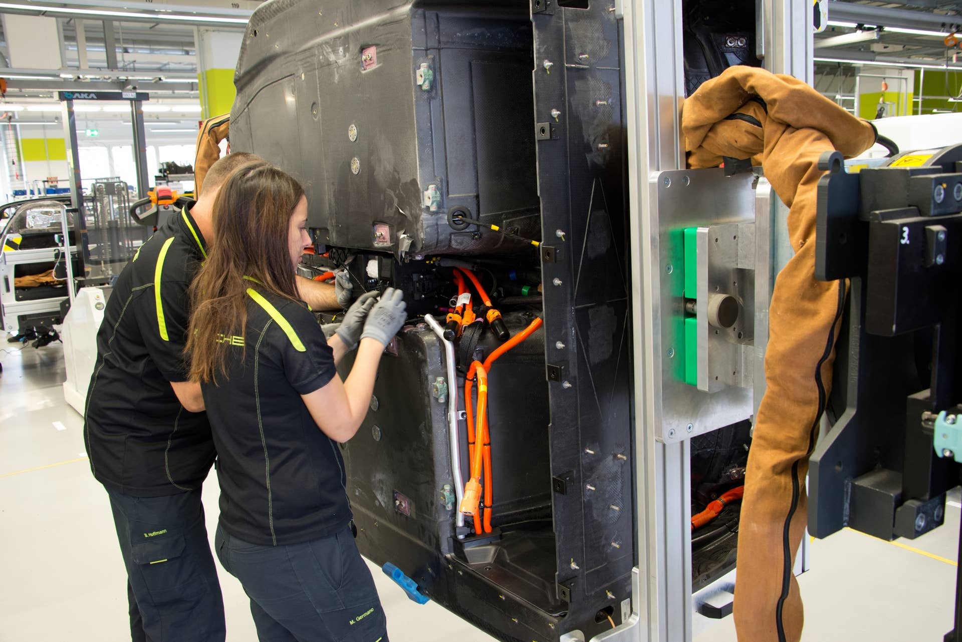 The carbon fiber reinforced plastic (CFP) monocoque passenger tub is mounted on a motorized arm that allows it to be rotated 360 degrees, and it can be raised or lowered to ease assembly. In this picture, two workers are installing the main wiring harness (orange) for the hybrid power system. An aluminum water line (silver), to cool the battery, is also visible. The large open space, to the right of the female worker, will be occupied by the primary drive battery.