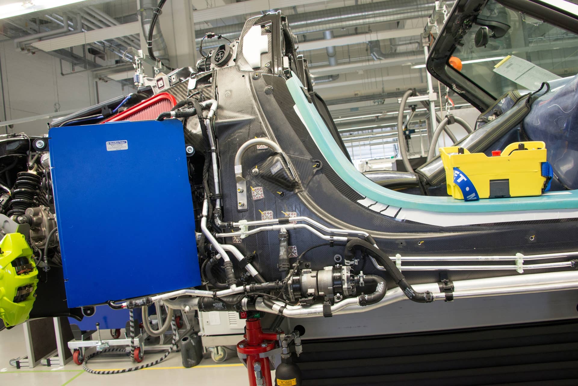 Still lacking nearly all of its body panels, the tub is fitted with the various pumps and liquid cooling lines that will serve to keep the engine, motors and battery pack within proper operating temperatures. The large blue panel, on the left side of the image, is a cover to protect the radiator from damage during assembly. The red item at the bottom, with the pressure cylinder next to it, is used to raise and lower the body to ease assembly.