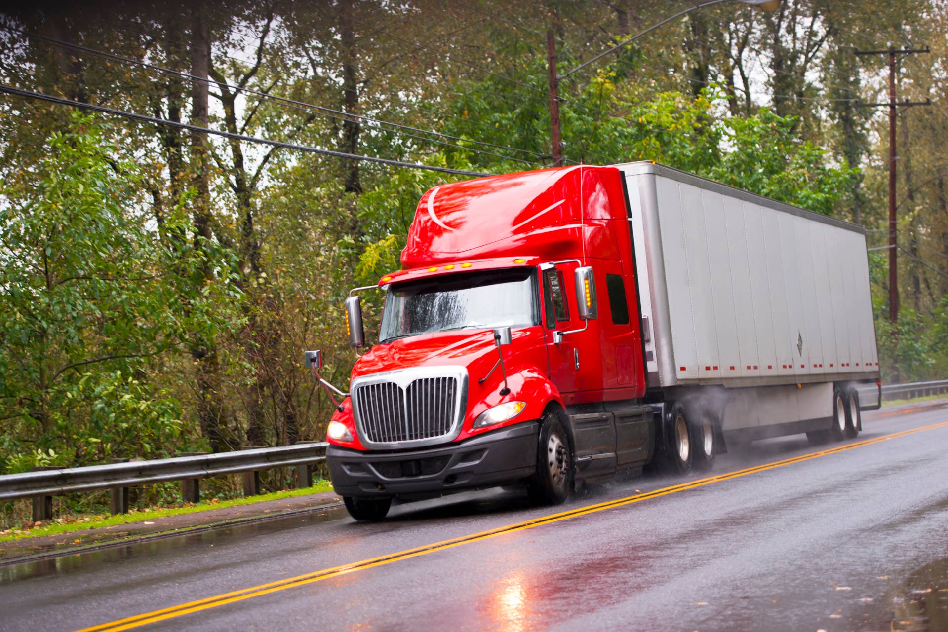 Big red semi truck shiny and wet from the rain with the reflection of light with a long distance measuring trailer with dust rain under the wheels and reflection of headlights on a wet road passing by green trees.