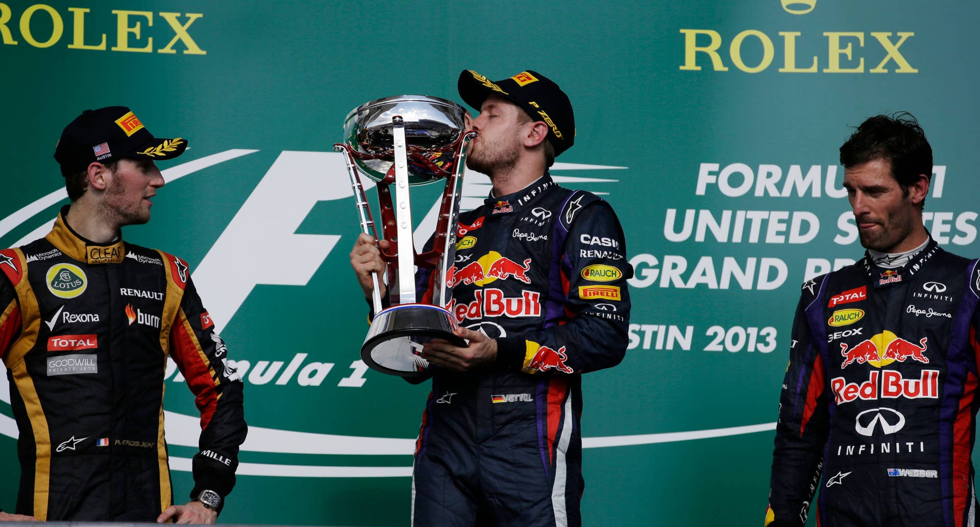 Second place finisher Lotus driver Romain Grosjean of Switzerland, left and second place finisher Red Bull driver Mark Webber of Australia watch as winner Red Bull driver Sebastian Vettel of Germany kisses his trophy after the Formula One U.S. Grand Prix auto race at the Circuit of the Americas, Sunday, Nov. 17, 2013, in Austin, Texas. (AP Photo/Darron Cummings)