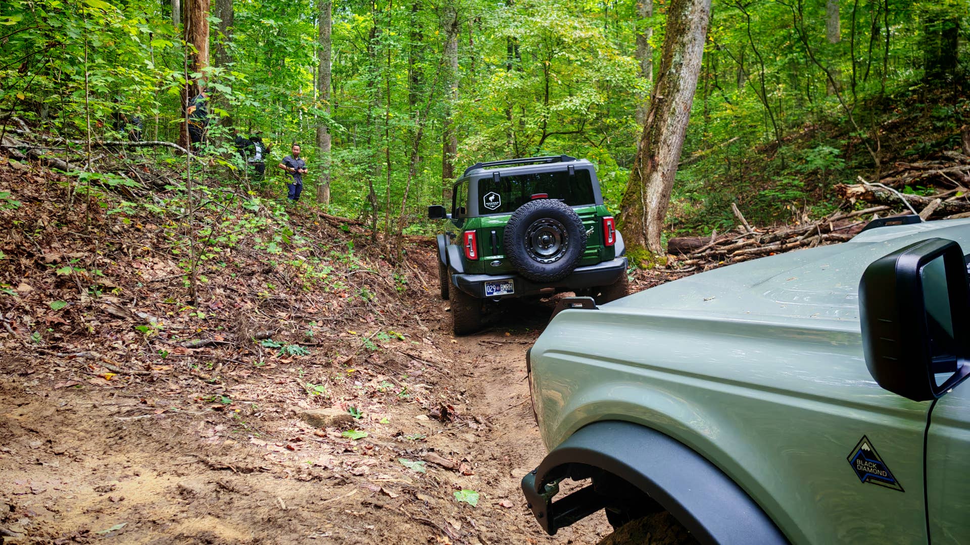 The front of a light green Ford Bronco as it follows a dark green Bronco through the woods