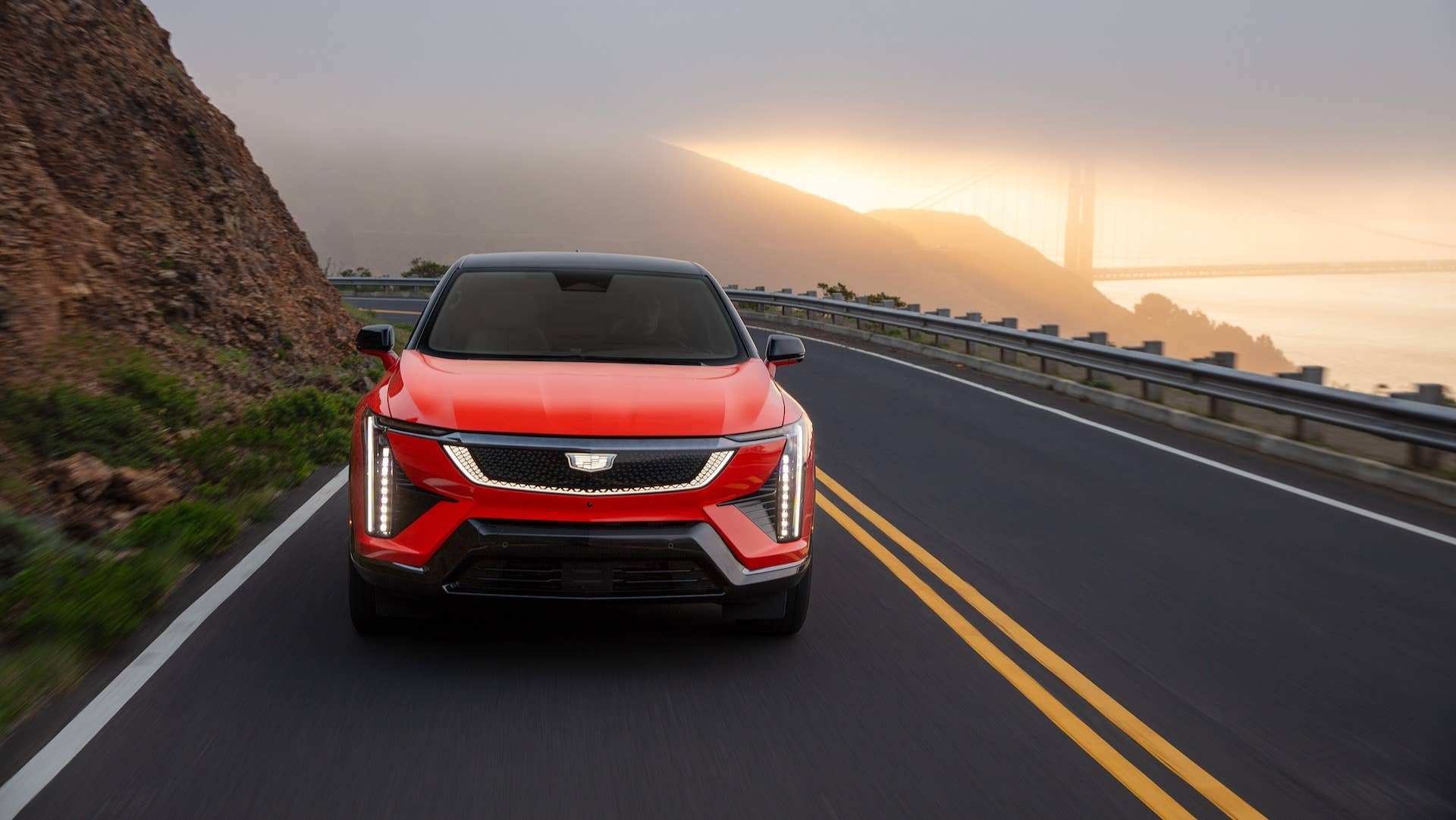 Front-facing view of the 2025 Cadillac OPTIQ in Monarch Orange driving on a mountain road with a scenic view in the background.
