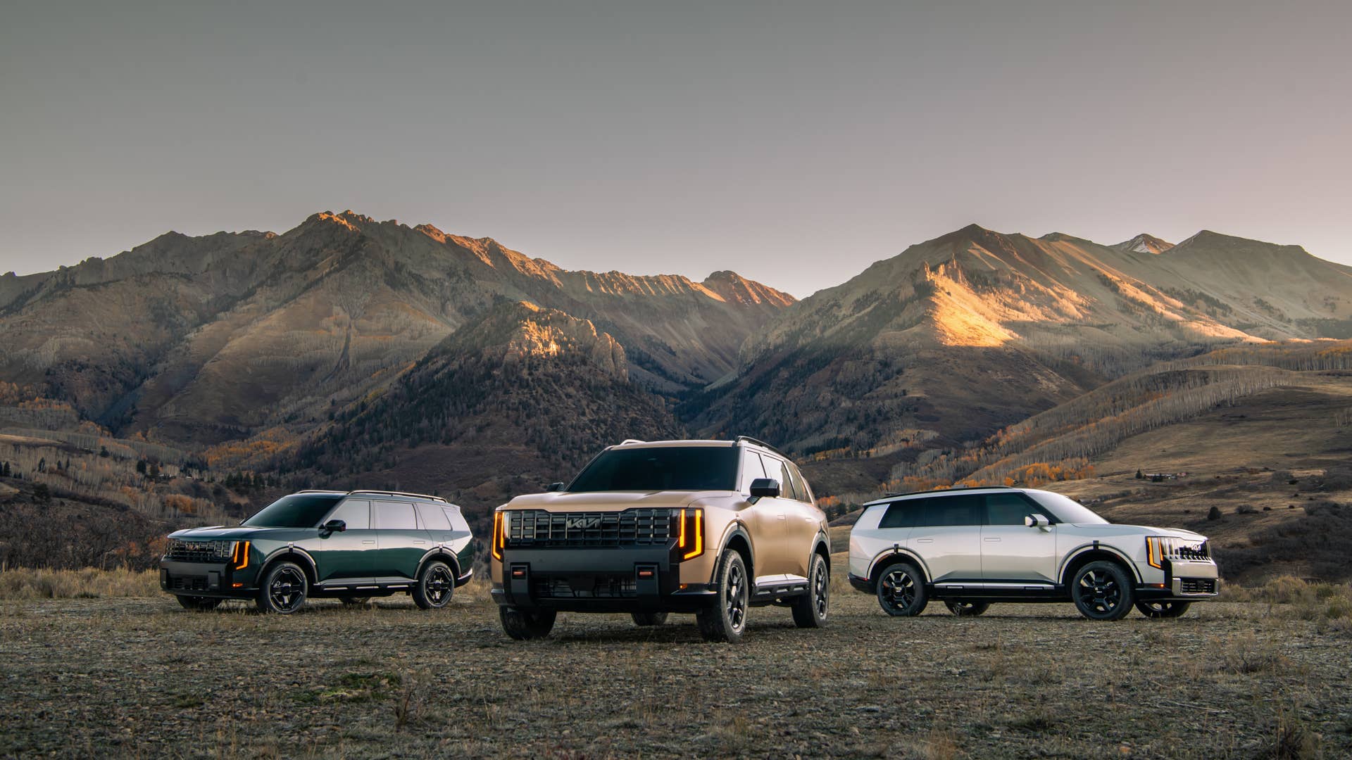 2027 Kia Telluride X-Pro in Terrain Brown flanked by two other Telluride trims parked on open ground in front of a mountain range at sunset