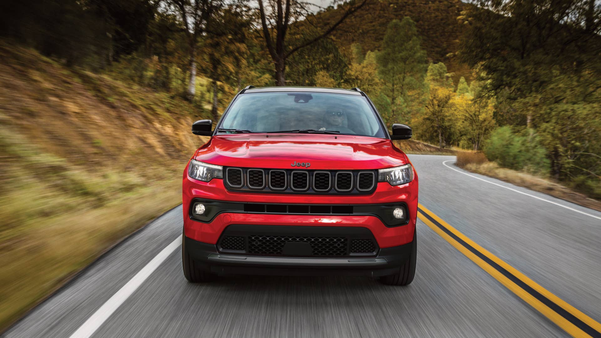 2027 Jeep® Compass Limited Altitude in Red Hot Pearl driving on tree lined road with cloudy skies and hills visible in the distance.